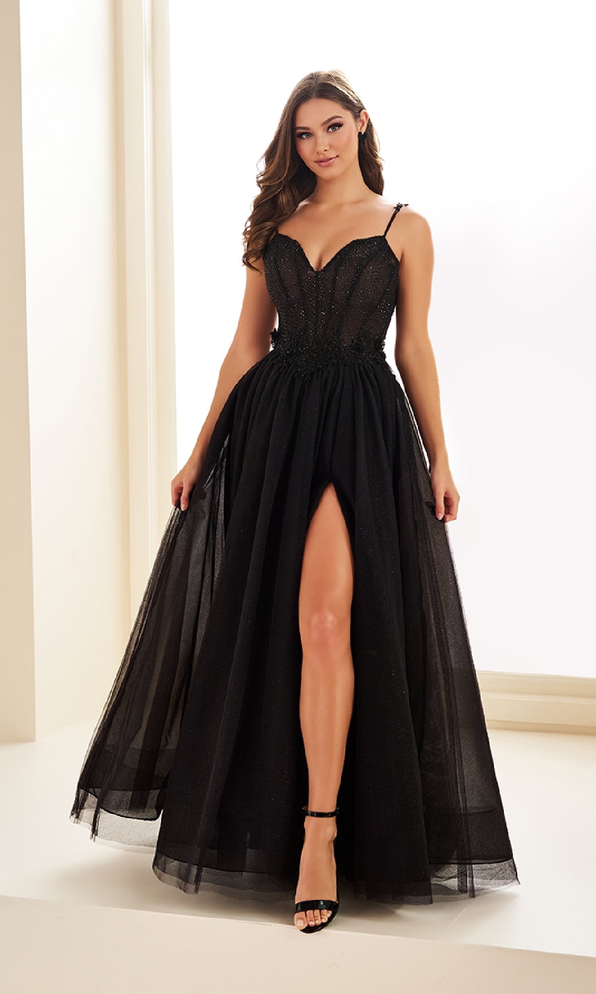 A woman wearing the Ellie Wilde EW36058 Long Prom Dress—a black, sleeveless gown with a thigh-high slit and floral lace embroidery—stands in a well-lit room, holding the sides of her dress.